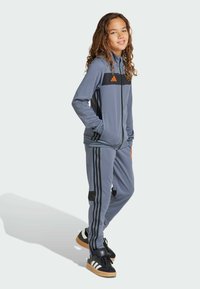 Chándal gris que presenta una chaqueta con cremallera con acentos en negro y naranja, y pantalones a juego con tres rayas negras en cada pierna.