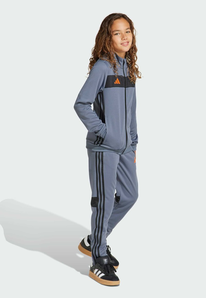 Chándal gris que presenta una chaqueta con cremallera con acentos en negro y naranja, y pantalones a juego con tres rayas negras en cada pierna.