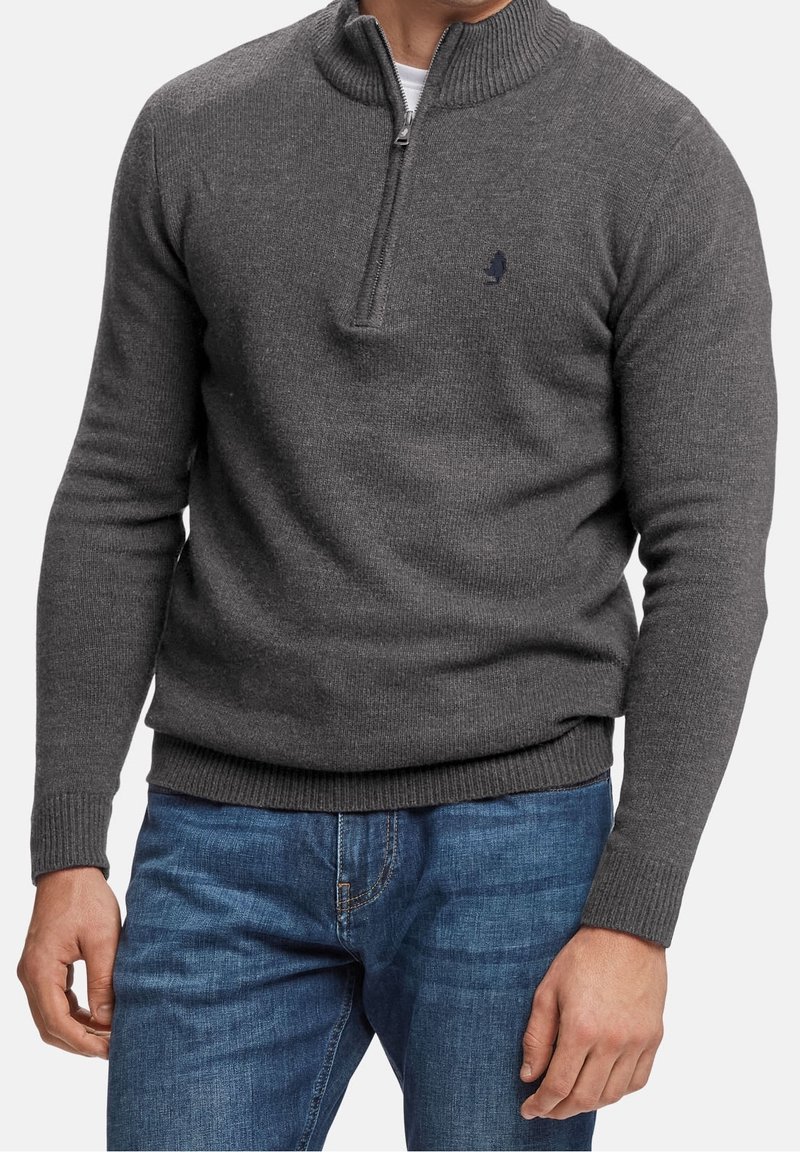 Homme portant un pull col zippé gris foncé et un jean bleu, se tenant devant un fond clair uniforme.
