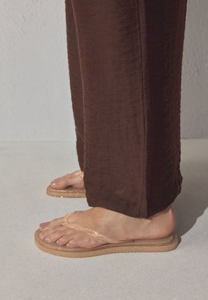 Pies con chanclas beige brillantes combinadas con pantalones anchos marrones, de pie sobre una superficie gris claro.