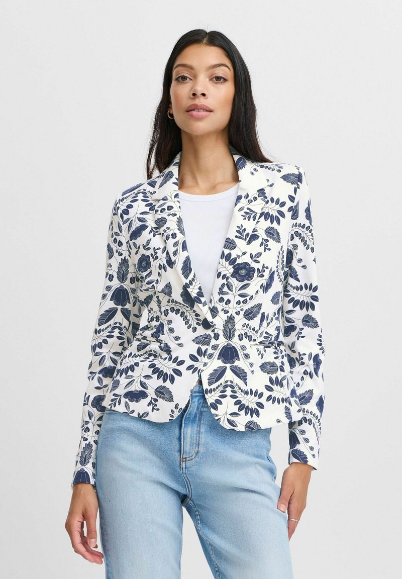 Blazer floreale bianco con motivi blu navy, revers a punta e tasche anteriori. Indossato sopra un top bianco e abbinato a jeans azzurri chiari.