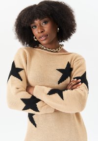 Knit sweater in beige with black star patterns on sleeves, crew neck, and ribbed cuffs. Soft texture and relaxed fit.
