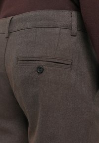 Brown tailored trousers made of textured fabric, featuring a back pocket with a black button. Neat stitching along the edges.
