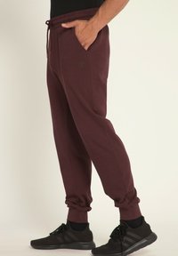 Bordowe dresy z elastycznym pasem i sznurkiem, z kieszeniami bocznymi i ściągaczami na kostkach, w połączeniu z czarnymi butami sportowymi.