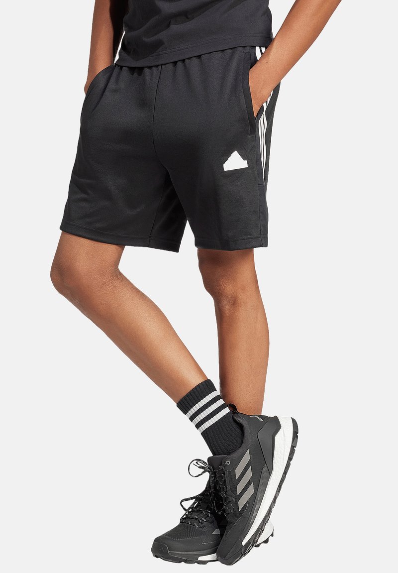 Schwarze Sportshorts mit elastischem Bund, seitlichen Taschen und weißen drei Streifen als Akzent. Getragen mit schwarzen Schuhen und gestreiften Socken.