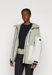 Kevyt, kaksivärinen takki harmaana ja valkoisena, jossa on teksturoitu kangas, säädettävä huppu, useita vetoketjuja ja elastiset hihansuut, käytetty mustan paidan päällä.