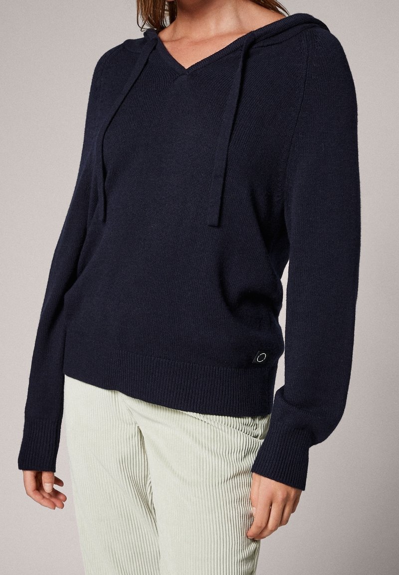 Personne portant un sweat à capuche en maille bleu marine foncé avec un col en V, des manches longues et un pantalon côtelé de couleur claire.