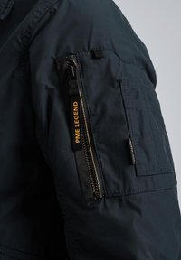 Marineblå bomberjakke med en lynlåslomme på venstre ærme, med sorte metaldele og "PME LEGEND" tekst i guld. Struktureret stof.