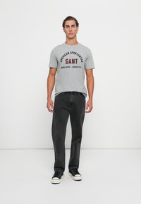 Grå bomulls t-skjorte med svart og rød GANT-logo tekst, kombinert med mørk denimjeans og svarte sneakers med hvite såler.