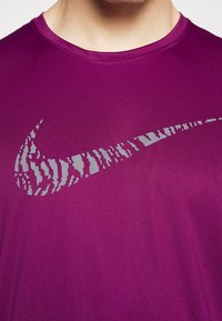 Lila Sport-T-Shirt mit einem großen, strukturierten, reflektierenden grauen Nike-Swoosh-Logo auf der Vorderseite, aus glattem, atmungsaktivem Material gefertigt.