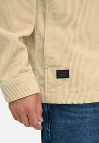 Hand met beige corduroy jas met zwarte stoffen label en blauwe spijkerbroek zichtbaar bij de taille.