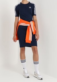 Mörkblå klänning med korta ärmar, vita tre-strimmiga detaljer, kombinerad med en orange tröja knuten i midjan; vita knästrumpor och sneakers.