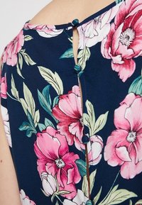 Navy kukka-paita, jossa on suuria vaaleanpunaisia ja valkoisia kukkia, takana napitus ja lyhyet hihat. Sileä kangas.