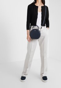 Borsa rotonda blu navy con manico ad anello e tracolla regolabile. Il modello indossa un cardigan nero, una maglietta bianca e pantaloni grigio chiaro con scarpe blu navy.