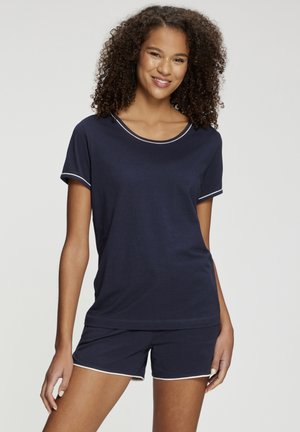 Smiling woman with curly hair wearing navy blue short-sleeve top and matching shorts with white trim, standing against a plain background.