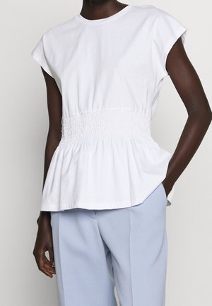Vrouw draagt een witte peplum top met korte mouwen en een aangesloten taille, gecombineerd met lichtblauwe broek, staand met één hand in de zak.