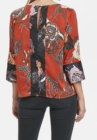 Blusa floreale rossa con dettagli neri, caratterizzata da maniche a tre quarti, uno scollo a chiave sul retro e un tessuto liscio con una finitura lucida.