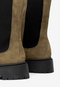 Botines de ante verde oliva con un panel lateral elástico negro y suela de goma negra resistente con texturas para un mejor agarre.