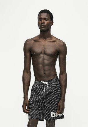 Man zonder shirt die frontaal staat, draagt zwarte zwemshorts met patroon en witte trekkoord op een effen witte achtergrond.
