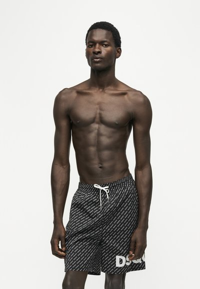 Homme torse nu debout face à l'avant portant un short de bain noir à motifs avec un cordon blanc sur fond blanc uni.