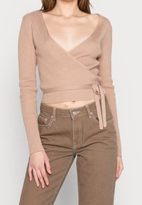 Haut wrap beige clair à côtes avec des manches longues, décolleté en V plongeant et un lien à la taille, associé à un pantalon marron taille haute avec poches.