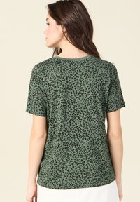 Camiseta verde de manga corta con un patrón de estampado de leopardo negro, escote redondo y un ajuste relajado. El material parece suave y transpirable.
