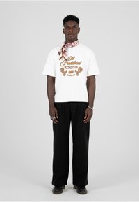 Valge ümar kaelusega T-särk pruun "Prohibited Social Club Berlin" graafikuga, kombineerituna lõdva musta pükste ja mustriga salliga.