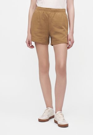 Person iført brune shorts med elastik i taljen og hvide sneakers med brune såler, stående mod en ensfarvet hvid baggrund.
