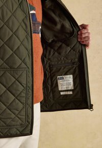 Person hält eine offene, dunkelgrüne gesteppte Jacke, die eine innere Reißverschlusstasche mit weißem Etikett zeigt.