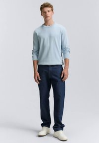 Világoskék kötött pulóver bordázott mandzsettákkal, sötétkék farmernadrággal és fehér tornacipőkkel, neutrális háttéren.