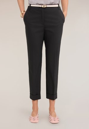 Femme portant un pantalon noir court avec ourlets retournés, une ceinture blanche avec boucle dorée, des bracelets dorés et des ballerines roses ornées.