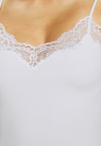 Camisole blanche avec un décolleté en V orné d'un délicat bord en dentelle blanche, mettant en valeur une texture lisse et douce. Bretelles spaghetti réglables.