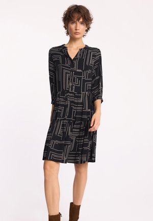 Kobieta w czarnej sukience do kolan z białymi geometrycznymi kropkami i rękawami do trzech ćwiartek, w połączeniu z brązowymi botkami za kostkę.