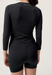 Persona che indossa un top aderente a maniche lunghe nero e pantaloni corti neri a vita alta, vista di spalle su uno sfondo chiaro e semplice.