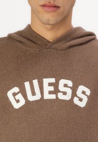 Felpa marrone realizzata in morbido tessuto con un piccolo logo bianco che riporta la scritta "GUESS" sulla parte anteriore. Presenta un cappuccio con cordini e orlo a costine.