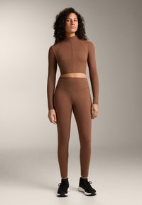 Brązowy strój sportowy składający się z krótkiego topu z długim rękawem i wysokim kołnierzem oraz pasujących legginsów z wysokim stanem, wykonany z gładkiego, elastycznego materiału.