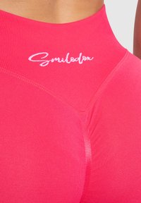 Top sportivo rosa aderente con un collo alto, materiale a maglia testurizzato, caratterizzato da un logo bianco e cuciture lisce lungo le spalle.