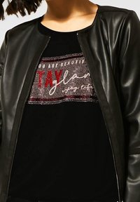 Černá bunda bomber z umělé kůže se zipovým zapínáním, nošená přes černé tričko s třpytivým nápisem z červených a bílých kamínků.