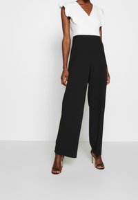 Svart jumpsuit med en åtsittande vit topp, som har volang-korta ärmar och vida byxor. Slätt tyg med en strukturerad design.