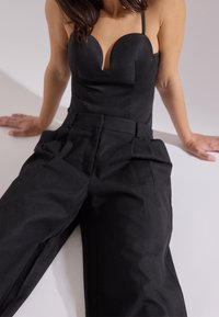 Donna seduta con le mani su una superficie, indossando un top nero senza spalline e pantaloni larghi neri a vita alta, su uno sfondo neutro.