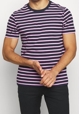 Man in een navyblauw en roze horizontaal gestreept T-shirt en zwarte broek, met zijn handen voor zich in elkaar geklapt tegen een effen achtergrond.