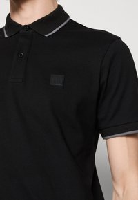 Homme portant un polo noir avec une bordure grise au col et aux manches, avec un petit logo embossé "BOSS" sur la poitrine.