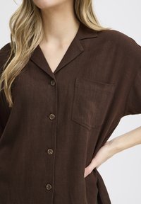 Chemise brune à manches courtes avec boutons, faite de tissu texturé. Inclut une poche poitrine et cinq boutons bruns sur le devant.