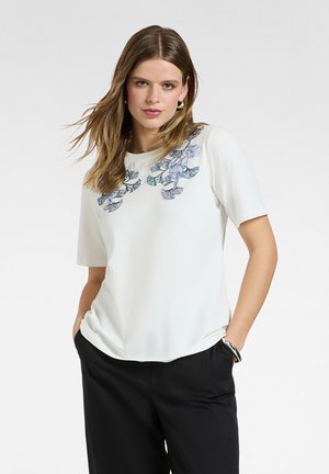 Mujer de cabello castaño claro viste una camiseta de manga corta blanca con bordados florales azules y pantalones negros, con las manos en los bolsillos, frente a un fondo liso.