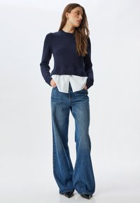 Pull à manches longues côtelé bleu marine superposé à une chemise blanche à col, associé à un jean bleu à jambe large et des chaussures noires à bout pointu.