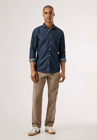 Mörk denimskjorta med knappar och uppkavlade ärmar, kombinerad med ljusbruna byxor och vita sneakers. Har en framficka och klassisk passform.