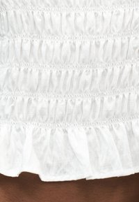 Λευκό ύφασμα με οριζόντιο ρελιασμένο τσαλάκωμα με λάστιχο και λεπτομέρεια ραμμένης βολάν στη βάση.