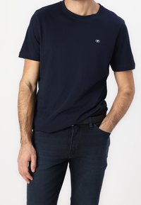 Navy blue cotton t-shirt with a round neckline, short sleeves, and a small logo on the chest, paired with dark denim jeans.