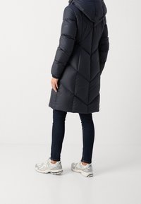 Abrigo acolchado navy con capucha, con costuras en forma de chevrón y puños elásticos, combinado con jeans de mezclilla oscuros y zapatillas deportivas plateadas.