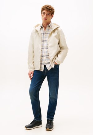 Lichtbeige jack met een capuchon en elastische manchetten, gedragen over een blauw-wit geruit overhemd. Donkerblauwe jeans en zwarte sneakers maken de outfit compleet.